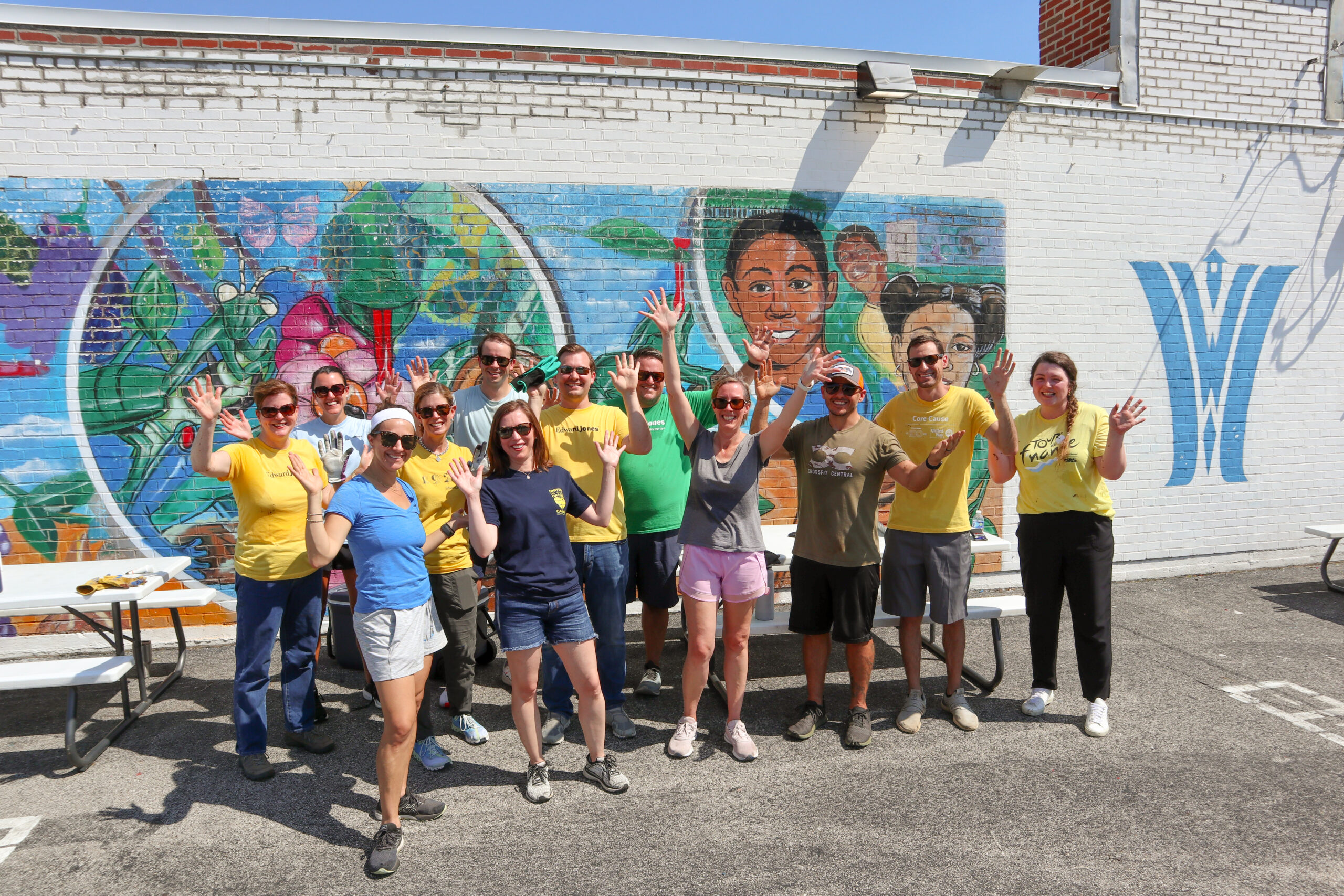 A group of people smiling and waving in front of a colorful mural on a sunny day. They stand on the pavement, wearing casual clothes, with picnic tables nearby. The mural features vibrant illustrations and a large face.
