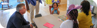 A man in a suit kneels beside a table where three children wearing colorful clothing and headbands are seated, smiling and playing a board game with an adult. This lively scene is set in a bright room with a colorful rug and open space in the background.